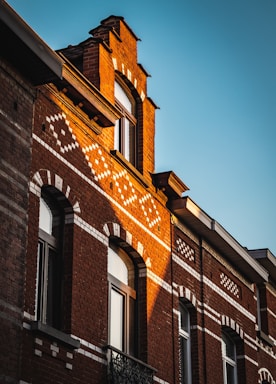 A building facade featuring intricate brickwork with decorative patterns and large windows. The sunlight casts warm tones on the red bricks, accentuating the architectural details.