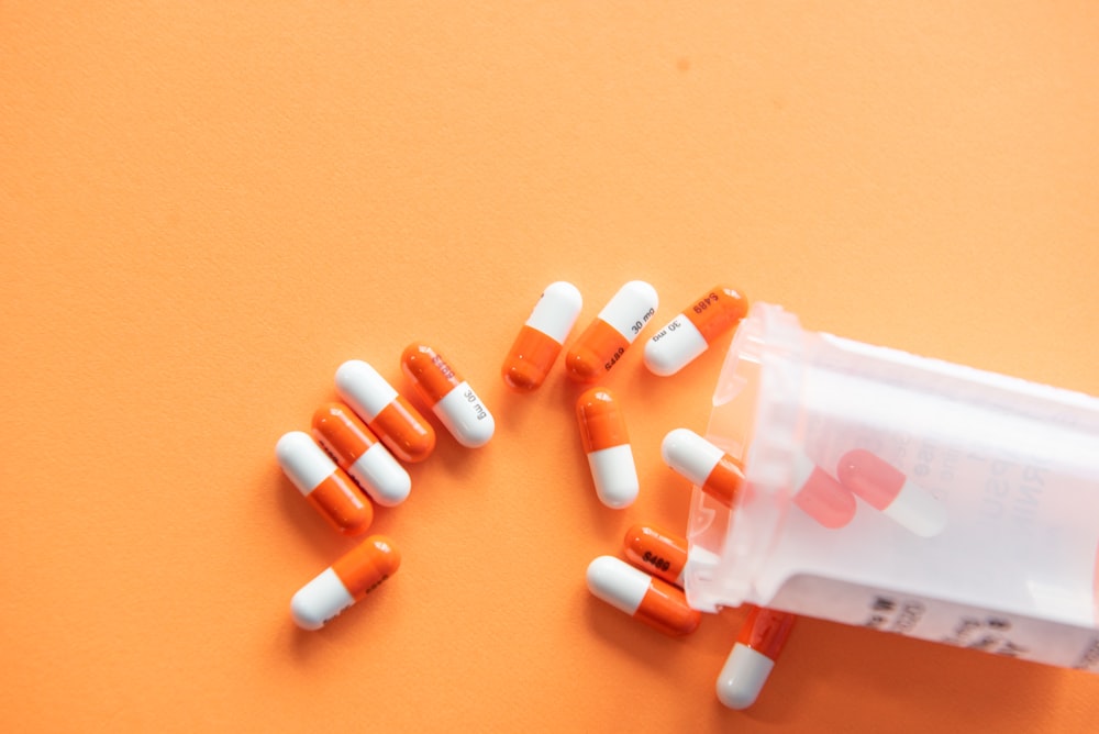 Orange background. There is a clear plastic bill bottle tipped on its side, and orange and white pills have come out of it. 