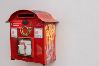 A red, vintage-style mailbox attached to a plain white wall, featuring some graffiti and stickers on its surface. The mailbox has a golden emblem and a small window displaying postal collection times.