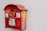 A red, vintage-style mailbox attached to a plain white wall, featuring some graffiti and stickers on its surface. The mailbox has a golden emblem and a small window displaying postal collection times.