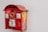 A red, vintage-style mailbox attached to a plain white wall, featuring some graffiti and stickers on its surface. The mailbox has a golden emblem and a small window displaying postal collection times.