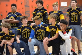 A group of young athletes wearing blue and yellow jerseys are gathered in a gym with climbing walls in the background. The jerseys have numbers and the words 'West Hernando' on them. Some are standing, while others are kneeling, showing camaraderie and team spirit.