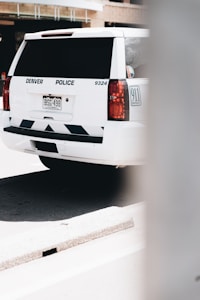 A white police SUV parked on a street, displaying 'Denver Police' on the rear. The word '911 Emergency' is visible along with its license plate. The background shows an urban environment and part of a concrete barrier is also visible.