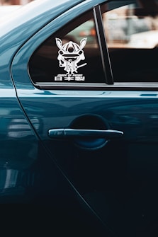 A side view of a car with a blue exterior, featuring a window with a decal of a stylized samurai helmet and Japanese text that reads 'Made in Japan'. The car door handle is visible below the window.