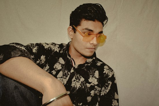 A person wearing a floral-patterned shirt and orange-tinted sunglasses leans slightly with a contemplative expression. The background is plain, emphasizing the subject's relaxed yet stylish appearance.