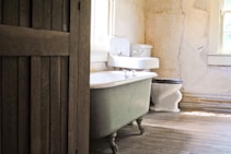 A rustic bathroom featuring a clawfoot bathtub, a vintage sink, and an old-fashioned toilet. The walls are aged, showing cracks and peeling paint, complemented by wooden floors with visible wear. Sunlight filters through a window, casting light on the wooden elements and creating a serene atmosphere.