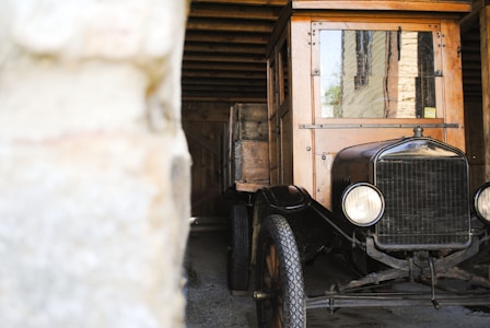 A vintage automobile with wooden and metal elements is parked in a rustic garage or barn. The vehicle has a classic design with circular headlights and a vintage-style front grille. The garage's wooden beams and stone walls create a historic and quaint atmosphere.