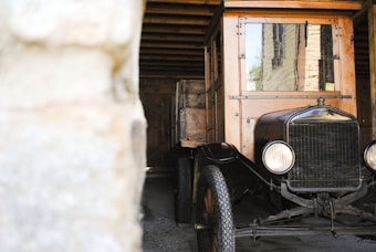 A vintage automobile with wooden and metal elements is parked in a rustic garage or barn. The vehicle has a classic design with circular headlights and a vintage-style front grille. The garage's wooden beams and stone walls create a historic and quaint atmosphere.