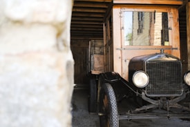 A vintage automobile with wooden and metal elements is parked in a rustic garage or barn. The vehicle has a classic design with circular headlights and a vintage-style front grille. The garage's wooden beams and stone walls create a historic and quaint atmosphere.
