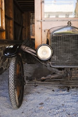 A vintage car with wooden and metal parts is parked inside a rustic shed. The car has a round headlamp, thin tires with distinct treads, and a front grille. The background includes wooden walls and a stone wall on the left side.