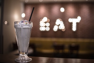 A tall glass filled with a creamy milkshake topped with colorful sprinkles and a black straw is prominently featured. The background is blurred, with illuminated letters spelling 'EAT' against a brick wall.