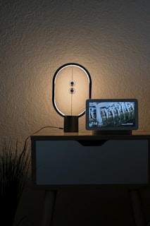 A modern lamp with a circular design is placed on a wooden nightstand. Next to it is a digital screen displaying a nature scene and the time, showing 7:14. There is a plant with long leaves at the side of the nightstand. The wall behind is textured in a neutral color.
