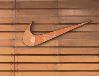 A large, prominent swoosh logo is mounted on a wooden slatted surface. The wood has a warm, brown tone, and the swoosh is a glossy orange color with subtle reflections and some text markings.