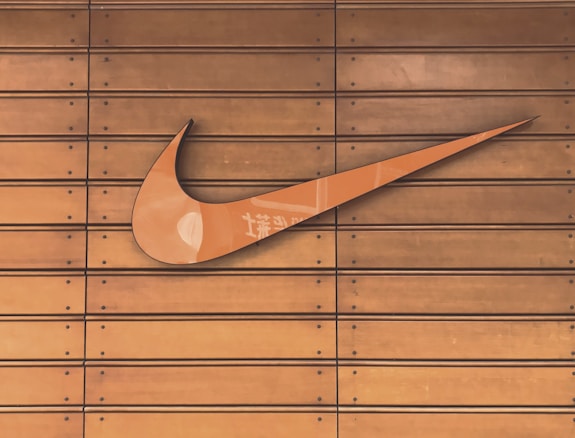 A large, prominent swoosh logo is mounted on a wooden slatted surface. The wood has a warm, brown tone, and the swoosh is a glossy orange color with subtle reflections and some text markings.
