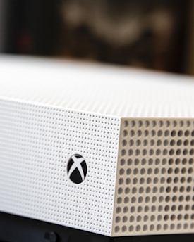 A close-up shot of a gaming console with a white textured surface featuring small circular perforations. The Xbox logo is visible on the side.