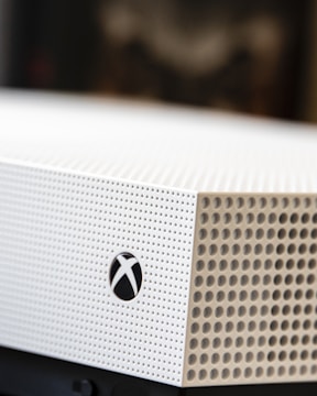 A close-up shot of a gaming console with a white textured surface featuring small circular perforations. The Xbox logo is visible on the side.