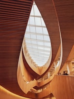 A modern architectural interior featuring curved wooden elements and a large glass ceiling allowing natural light to illuminate the space. The design creates a sense of flow and openness with multiple levels visible and people standing along the railing.