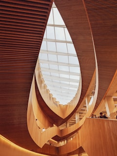 A modern architectural interior featuring curved wooden elements and a large glass ceiling allowing natural light to illuminate the space. The design creates a sense of flow and openness with multiple levels visible and people standing along the railing.