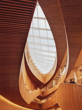 A modern architectural interior featuring curved wooden elements and a large glass ceiling allowing natural light to illuminate the space. The design creates a sense of flow and openness with multiple levels visible and people standing along the railing.