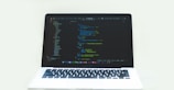 A laptop displaying a code editor with a dark theme, showing lines of colorful code. The environment is bright and minimalistic.