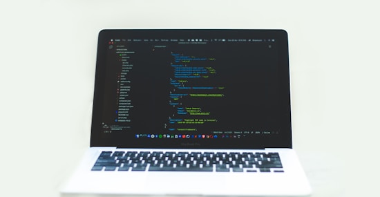 A laptop displaying a code editor with a dark theme, showing lines of colorful code. The environment is bright and minimalistic.