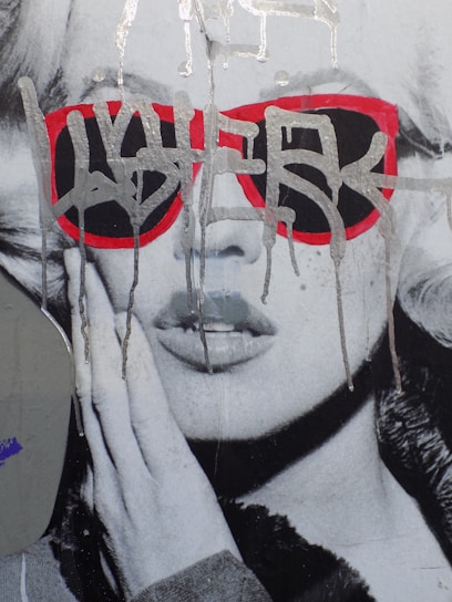 A monochrome close-up portrait of a woman wearing red sunglasses overlaid with drips of silver paint. The image is stylized and resembles street art or graffiti, giving it an urban, edgy feel.