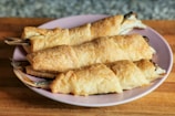 Three puff pastry-wrapped fish are presented on a light pink plate. The pastries are golden brown and flaky, showing a crisp texture. The fish tails are visible at the ends, indicating the fish was baked whole within the pastry.