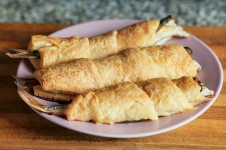 Three puff pastry-wrapped fish are presented on a light pink plate. The pastries are golden brown and flaky, showing a crisp texture. The fish tails are visible at the ends, indicating the fish was baked whole within the pastry.