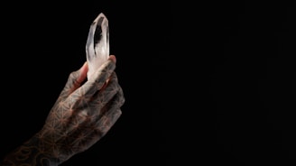 A tattooed hand holds a clear, multifaceted crystal against a dark background. The hand features intricate geometric patterns, and the crystal is illuminated, creating a striking contrast with the surrounding darkness.