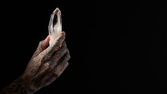 A tattooed hand holds a clear, multifaceted crystal against a dark background. The hand features intricate geometric patterns, and the crystal is illuminated, creating a striking contrast with the surrounding darkness.