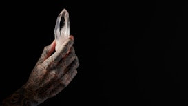 A tattooed hand holds a clear, multifaceted crystal against a dark background. The hand features intricate geometric patterns, and the crystal is illuminated, creating a striking contrast with the surrounding darkness.
