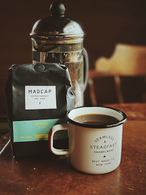 A French press coffee maker sits on a wooden table alongside a black bag of coffee labeled 'MADCAP Coffee Company'. In front of the bag is a white enamel mug filled with coffee, featuring text stating 'Seamless & Steadfast Enamelware' from 'Best Made Co. New York'. The warm tones create a cozy and inviting atmosphere.