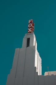 A tall, white Art Deco-style tower with geometric design elements, featuring a red sign displaying the word 'FOX' near the top of the structure. The sky is a clear, deep teal color, providing a striking contrast to the pale building.