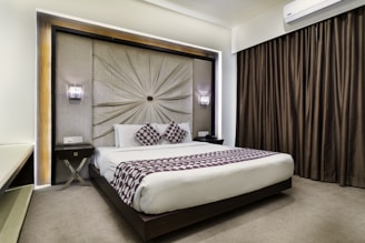 A modern bedroom featuring a large bed with white linens and two decorative pillows with a geometric pattern. The headboard is ornate with radial pleats and flanked by two wall sconces providing ambient lighting. Dark curtains cover the window, and a sleek air conditioning unit is mounted above. The room has a neutral color palette.