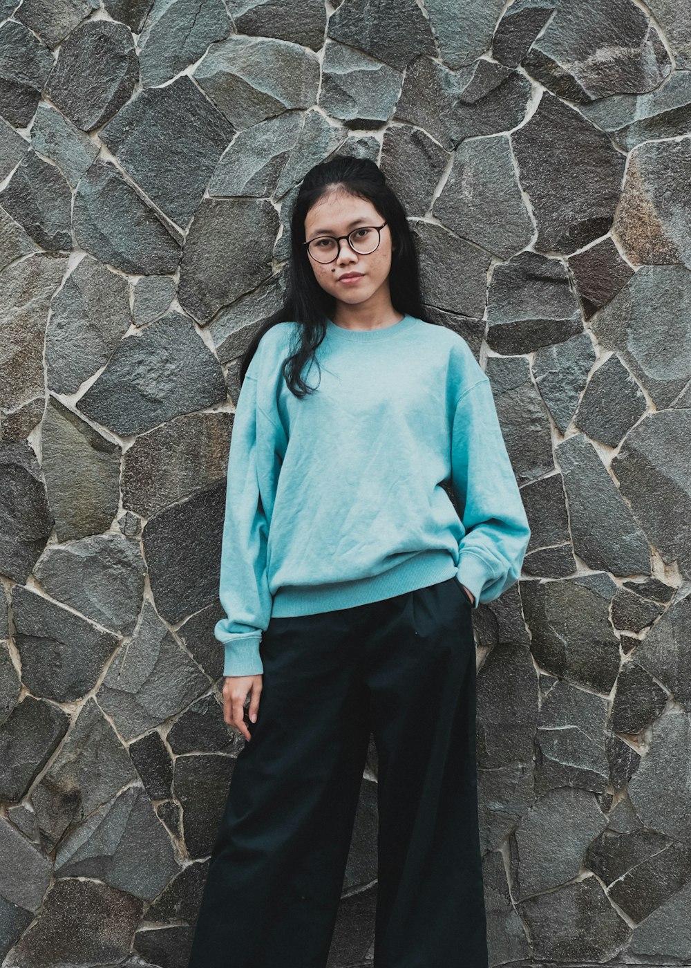 woman in teal long sleeve shirt and black pants standing beside gray wall