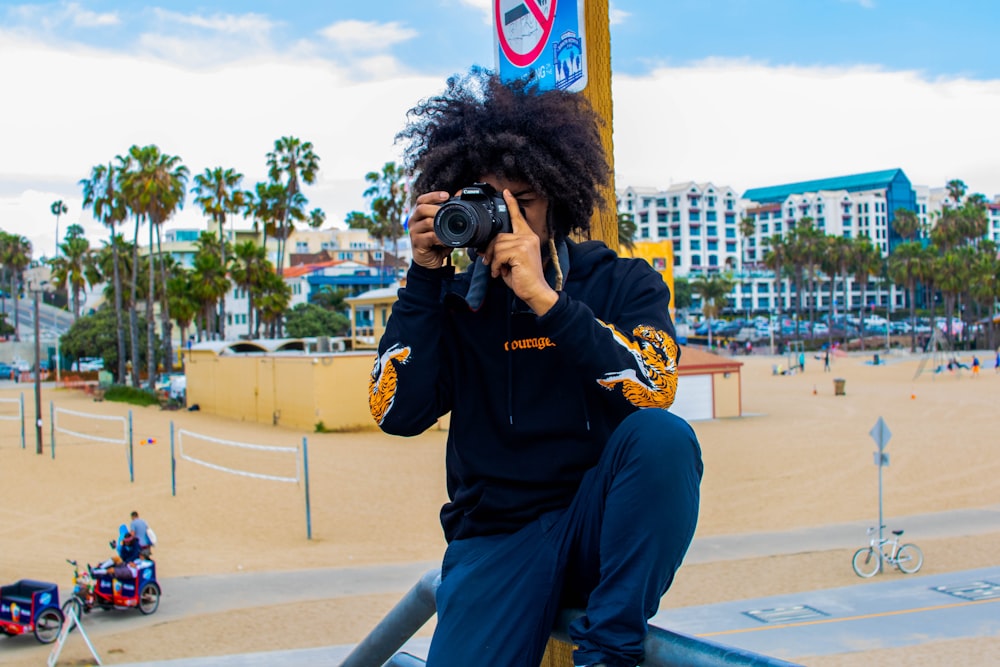 man in black jacket and blue denim jeans sitting on bench using black dslr camera