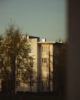 A residential building with a modern architectural design, featuring white walls and large windows. Trees with sparse foliage are in the foreground, and the scene is illuminated by warm, soft lighting, suggesting late afternoon or early evening.