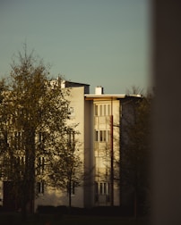A residential building with a modern architectural design, featuring white walls and large windows. Trees with sparse foliage are in the foreground, and the scene is illuminated by warm, soft lighting, suggesting late afternoon or early evening.