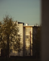 A residential building with a modern architectural design, featuring white walls and large windows. Trees with sparse foliage are in the foreground, and the scene is illuminated by warm, soft lighting, suggesting late afternoon or early evening.