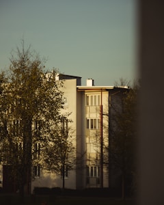 A residential building with a modern architectural design, featuring white walls and large windows. Trees with sparse foliage are in the foreground, and the scene is illuminated by warm, soft lighting, suggesting late afternoon or early evening.