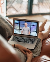 A person is sitting on a couch with a laptop on their lap, watching a video of a speaker standing in front of a colorful background. The scene is set indoors with a blurred background, and the laptop shows a paused or live video of the speaker.