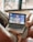 A person is sitting on a couch with a laptop on their lap, watching a video of a speaker standing in front of a colorful background. The scene is set indoors with a blurred background, and the laptop shows a paused or live video of the speaker.