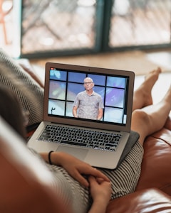 A person is sitting on a couch with a laptop on their lap, watching a video of a speaker standing in front of a colorful background. The scene is set indoors with a blurred background, and the laptop shows a paused or live video of the speaker.