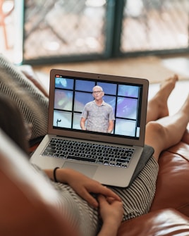 A person is sitting on a couch with a laptop on their lap, watching a video of a speaker standing in front of a colorful background. The scene is set indoors with a blurred background, and the laptop shows a paused or live video of the speaker.