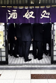 A group of people in suits is seen standing closely together under a traditional Japanese noren curtain with calligraphy. The ground is tiled, and the atmosphere suggests a business meeting or social gathering in a small space.