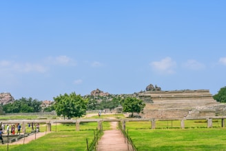 A historical site featuring ancient stone structures surrounded by lush greenery under a clear blue sky. Groups of people walk along a path leading to the ruins, with rocky hills visible in the background.