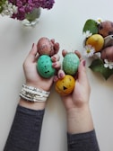 Hands holding several decorated Easter eggs with different facial expressions and words, alongside a small basket filled with similar eggs and flowers. A vase of purple and white flowers is visible in the background. The eggs are colorfully painted and adorned with simple drawings and text.