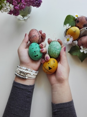 Hands holding several decorated Easter eggs with different facial expressions and words, alongside a small basket filled with similar eggs and flowers. A vase of purple and white flowers is visible in the background. The eggs are colorfully painted and adorned with simple drawings and text.