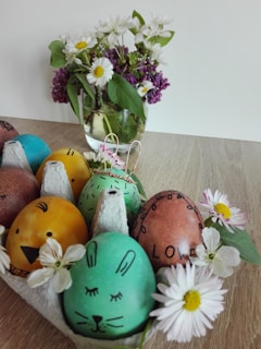 A wooden surface features a carton of colorfully decorated Easter eggs with faces, including a yellow one resembling a chick and a green one resembling a rabbit. Surrounding the carton are white daisies with yellow centers. In the background, a clear glass vase holds a bouquet of white and purple flowers with green leaves.
