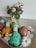 A wooden surface features a carton of colorfully decorated Easter eggs with faces, including a yellow one resembling a chick and a green one resembling a rabbit. Surrounding the carton are white daisies with yellow centers. In the background, a clear glass vase holds a bouquet of white and purple flowers with green leaves.